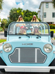 Group of friends laughing in a light blue MOKE beach buggy with a 'Walton County' sign, palm trees and coastal homes in the background.