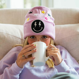 Cozy indoor shot of a person on a beige couch wearing a pastel pink beanie with embroidered stars and a black lightning-smiley patch, sipping from a white mug while wearing a lavender sweatshirt.