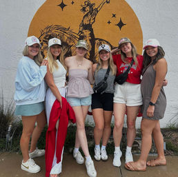Six friends in casual summer outfits and caps smiling and posing on a sidewalk in front of a large western-themed street mural of a cowboy on horseback on a white stucco wall.