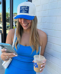 Smiling person in a blue tank top and white-and-blue trucker hat with a fish patch, standing against a white brick wall on a sunny street, holding a smartphone and an iced coffee in a clear cup with a blue flower sticker.