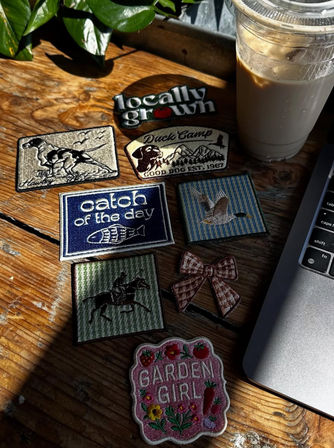 Sunlit wooden table with assorted embroidered patches (Garden Girl, catch of the day, duck, hunting), a gingham bow, iced coffee cup and laptop edge.