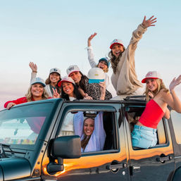 Group of friends in a black Jeep at a beach sunset, women in casual clothes and trucker hats laughing, waving and enjoying the coastal golden hour.