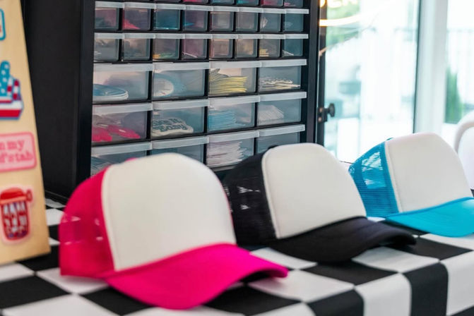 Colorful mesh trucker hats (pink, black, turquoise) with blank white fronts lined up on a black-and-white checkered merch table, with a plastic drawer organizer of stickers and patches in the background.