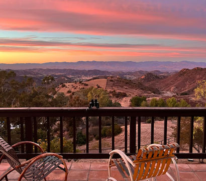 Serene sunset mountain view from a tiled patio — two vintage rocking chairs and binoculars on the railing overlooking rolling hills under a pink-purple sky.