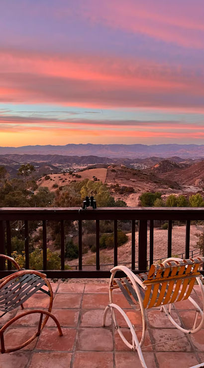 Serene sunset mountain view from a tiled patio — two vintage rocking chairs and binoculars on the railing overlooking rolling hills under a pink-purple sky.
