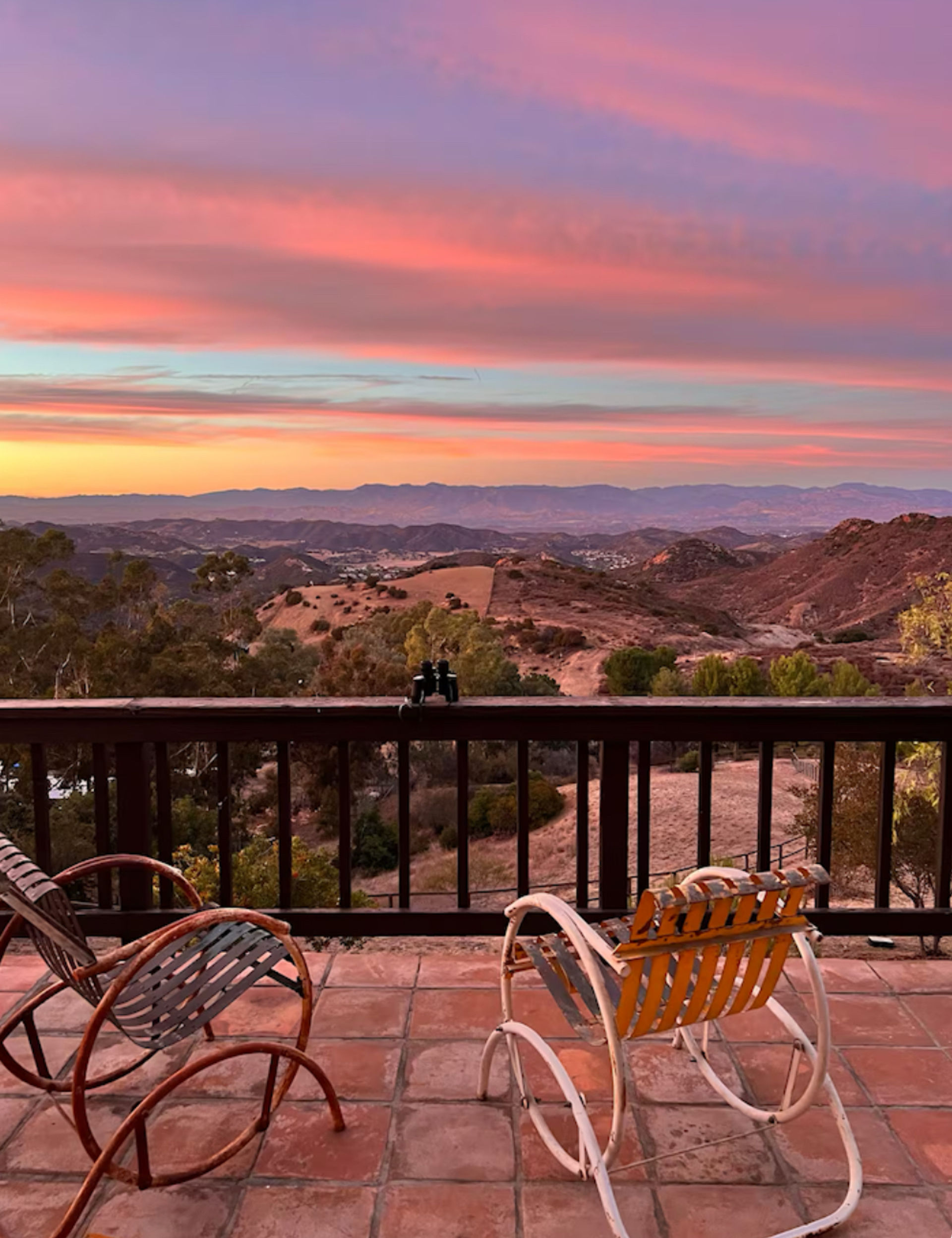 Serene sunset mountain view from a tiled patio — two vintage rocking chairs and binoculars on the railing overlooking rolling hills under a pink-purple sky.