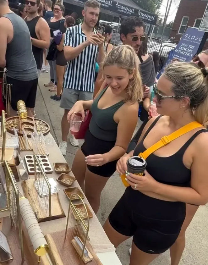 Two women browsing gold jewelry and trinkets at an outdoor street market stall, holding drinks amid a lively summer crowd