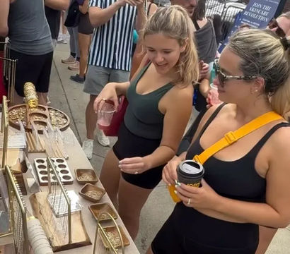 Two women browsing gold jewelry and trinkets at an outdoor street market stall, holding drinks amid a lively summer crowd