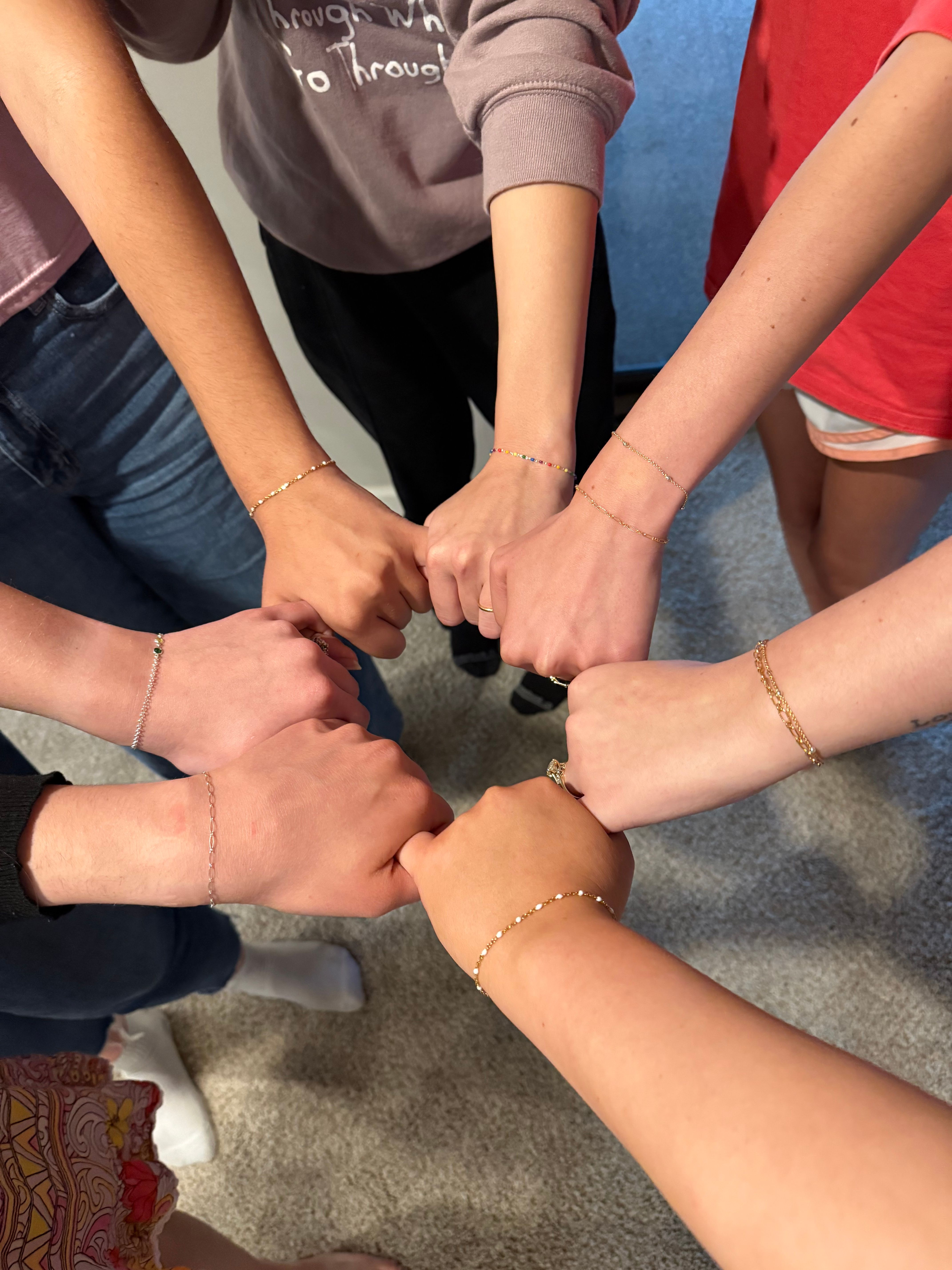 Overhead shot of a group joining fists in a circle, showcasing delicate gold and beaded bracelets on wrists against a carpeted floor — playful group wrist photo.