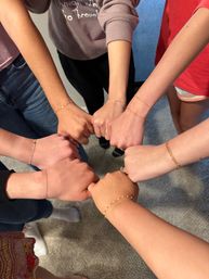 Overhead shot of a group joining fists in a circle, showcasing delicate gold and beaded bracelets on wrists against a carpeted floor — playful group wrist photo.