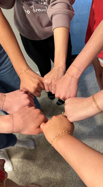 Overhead shot of a group joining fists in a circle, showcasing delicate gold and beaded bracelets on wrists against a carpeted floor — playful group wrist photo.