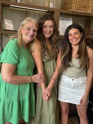 Three smiling women in green outfits posing together inside a cozy jewelry boutique with hanging necklaces and wicker display shelves