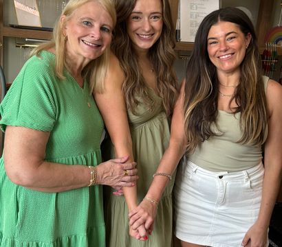 Three smiling women in green outfits posing together inside a cozy jewelry boutique with hanging necklaces and wicker display shelves