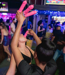 Crowded nightclub scene with two women taking shots from pink novelty penis-shaped shot dispensers under colorful bar lights, playful bachelorette-style party vibe