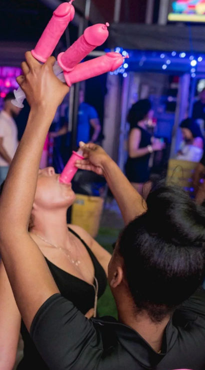 Crowded nightclub scene with two women taking shots from pink novelty penis-shaped shot dispensers under colorful bar lights, playful bachelorette-style party vibe