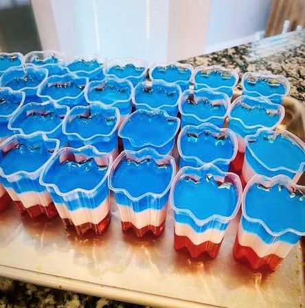 Rows of red, white, and blue layered gelatin desserts in clear flower-shaped cups on a kitchen countertop — festive USA Fourth of July jello cups.