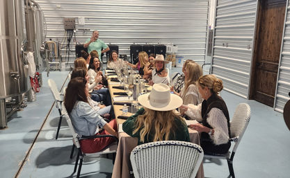 Cheerful group enjoying a private tasting dinner at a long communal table inside an industrial brewery-style space with stainless steel tanks and corrugated metal walls.