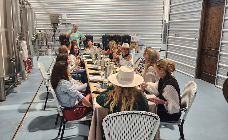 Cheerful group enjoying a private tasting dinner at a long communal table inside an industrial brewery-style space with stainless steel tanks and corrugated metal walls.