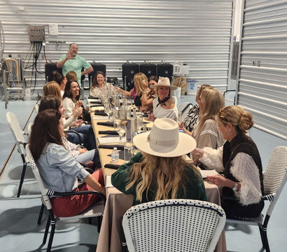 Cheerful group enjoying a private tasting dinner at a long communal table inside an industrial brewery-style space with stainless steel tanks and corrugated metal walls.
