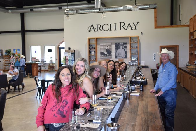 Smiling friends gathered at a long marble wine-tasting bar in a bright modern tasting room, sipping glasses while a man in a cowboy hat stands at the far end.