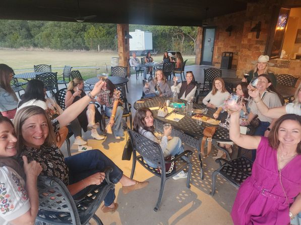 Group of friends toasting with wine glasses on a rustic covered patio at a countryside wine-tasting area, metal tables and grassy lawn beyond.