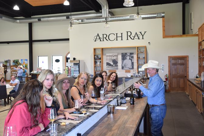 Winery tasting room interior with women smiling and sipping at a long marble tasting bar while a man in a cowboy hat pours wine from a bottle.