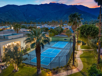 Aerial evening view of bright blue tennis courts at a palm-tree lined resort complex with winding paths and warm lights, set against rugged mountain foothills at sunset