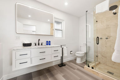 Bright modern white residential bathroom with wall-mounted floating vanity, marble countertop, matte-black faucet and hardware, LED-framed mirror, glass-enclosed stone shower with rain showerhead, white toilet, and light wood-look flooring.