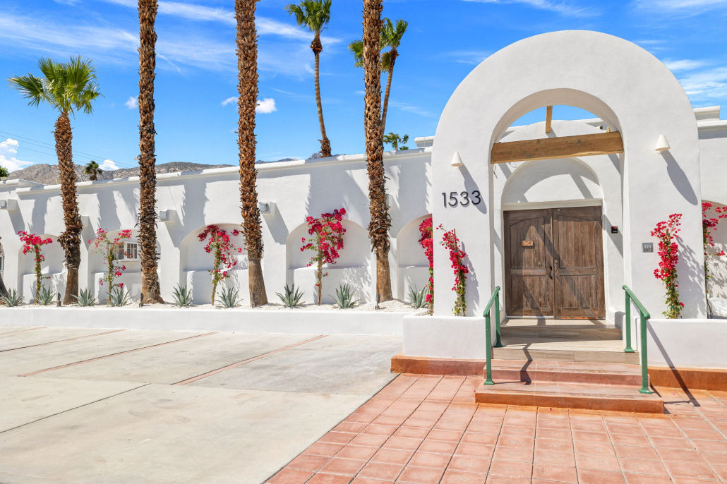 Sunlit Southwestern white stucco building with large arched entry and rustic wooden double doors, flanked by pink bougainvillea and tall palm trees, terracotta steps and concrete parking under a bright blue sky