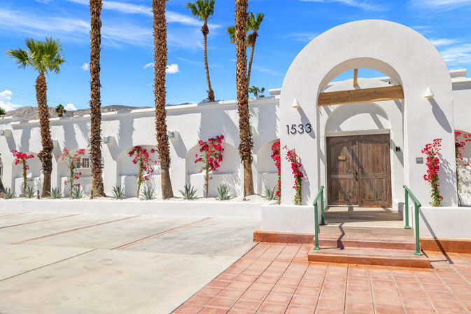 Sunlit Southwestern white stucco building with large arched entry and rustic wooden double doors, flanked by pink bougainvillea and tall palm trees, terracotta steps and concrete parking under a bright blue sky