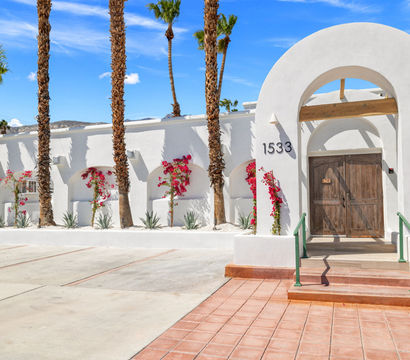 Sunlit Southwestern white stucco building with large arched entry and rustic wooden double doors, flanked by pink bougainvillea and tall palm trees, terracotta steps and concrete parking under a bright blue sky