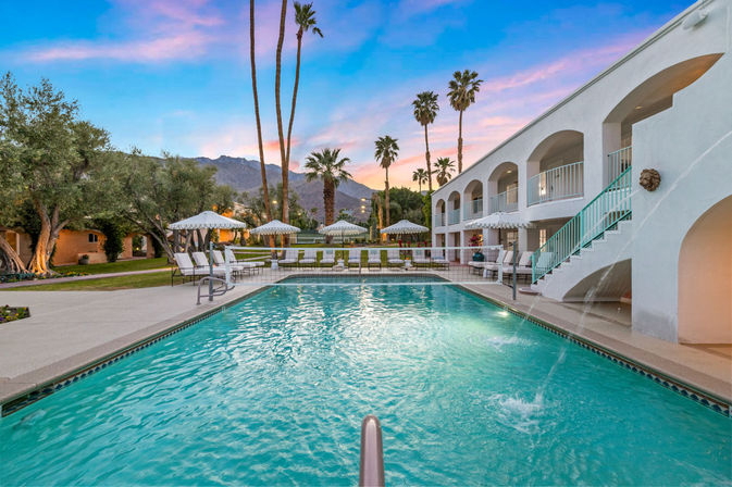 Turquoise outdoor pool at a white two‑story resort with arched balconies and teal stairs, lined with lounge chairs and umbrellas, tall palm trees and mountain backdrop at a pink‑blue sunset.