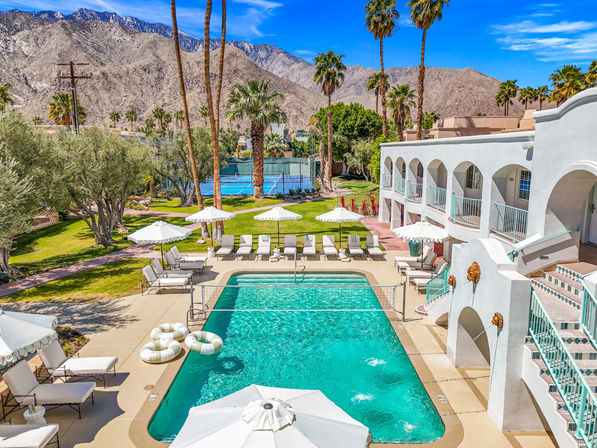 Sunny Palm Springs poolside: turquoise rectangular resort pool with white umbrellas and lounge chairs, palm trees, tennis courts and rugged desert mountains