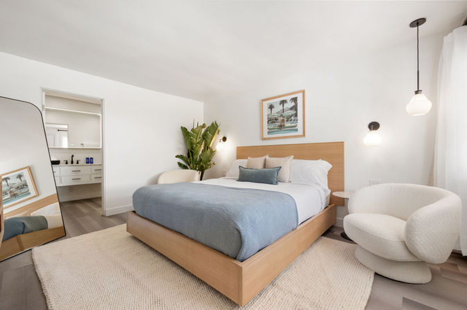 Airy coastal-style master bedroom with light wood platform bed dressed in blue coverlet and white linens, textured boucle swivel chair, tall potted plant, pendant lights, leaning full-length mirror and a glimpse of the en-suite bathroom.
