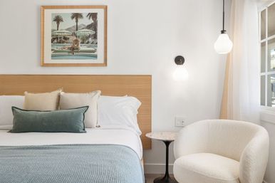 Bright modern bedroom with wood headboard, white linens, blue knit throw and green lumbar pillow, framed pool-and-lion artwork above bed, terrazzo side table, cream boucle armchair and hanging pendant lights by a sunlit window.