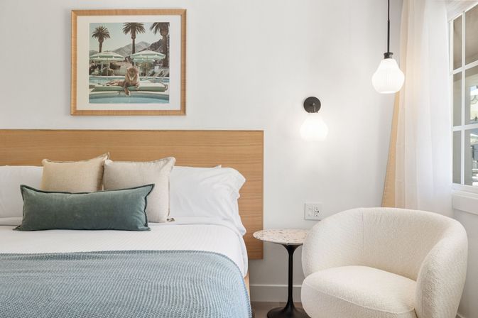 Bright modern bedroom with wood headboard, white linens, blue knit throw and green lumbar pillow, framed pool-and-lion artwork above bed, terrazzo side table, cream boucle armchair and hanging pendant lights by a sunlit window.