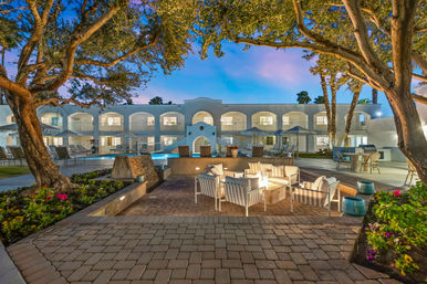 Twilight hotel courtyard with a glowing fire pit surrounded by striped lounge chairs, a lit pool, palm trees and arched white balconies.