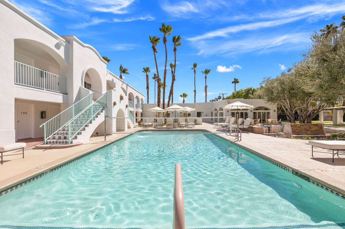 Sun‑soaked resort pool framed by white stucco arches, mint‑green staircase, lounge chairs and umbrellas, and tall palm trees against a bright blue sky.