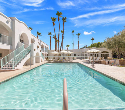 Sun‑soaked resort pool framed by white stucco arches, mint‑green staircase, lounge chairs and umbrellas, and tall palm trees against a bright blue sky.
