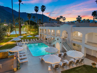 Sunset view of a white Mediterranean-style hotel courtyard with an illuminated rectangular pool, lounge chairs and scalloped umbrellas, palm trees and a desert mountain backdrop.