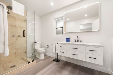 Bright modern residential bathroom with glass walk-in shower and beige tile, floating white vanity with marble countertop and illuminated LED mirror, matte black fixtures, toilet, and towel stand on light wood-look flooring.