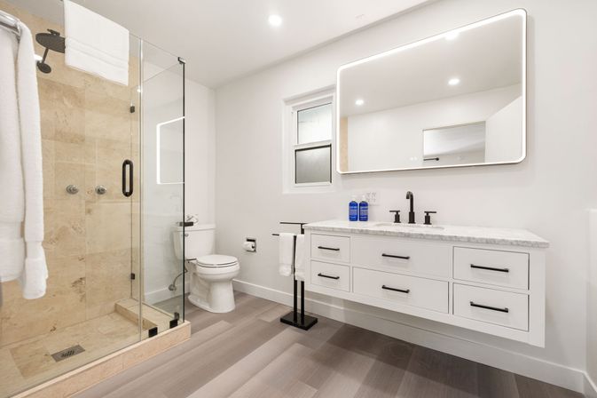 Bright modern residential bathroom with glass walk-in shower and beige tile, floating white vanity with marble countertop and illuminated LED mirror, matte black fixtures, toilet, and towel stand on light wood-look flooring.