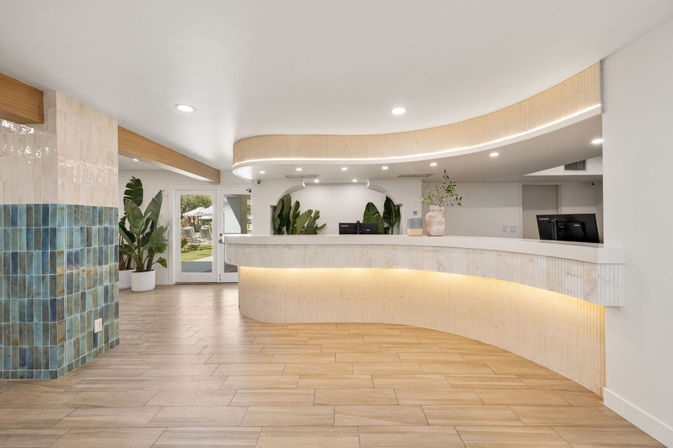 Bright modern hotel lobby reception with curved white desk and warm underlighting, wood-look tile floor, blue-green tiled column, potted tropical plants and glass doors to an outdoor patio