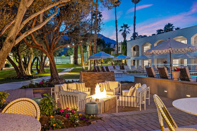 Twilight poolside resort courtyard with a glowing fire pit surrounded by striped lounge chairs, scalloped umbrellas, palm trees and a distant mountain backdrop.