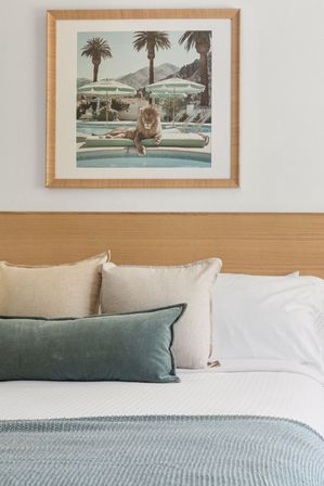 Cozy modern hotel bedroom with white linens, wooden headboard, beige and teal accent pillows, seafoam knit throw, and a framed tropical pool print of a lounging lion.