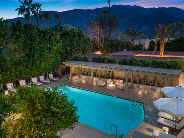 Twilight outdoor pool with glowing turquoise water, covered cabana with cushioned chairs and a fire pit, lounge chairs and umbrellas, framed by palm trees and rugged mountains.