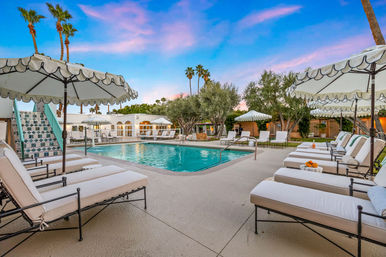 Sunset-lit desert resort pool with turquoise water, cushioned lounge chairs and scalloped umbrellas, tiled pool staircase, palm trees and manicured landscaping