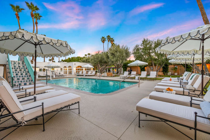 Sunset-lit desert resort pool with turquoise water, cushioned lounge chairs and scalloped umbrellas, tiled pool staircase, palm trees and manicured landscaping