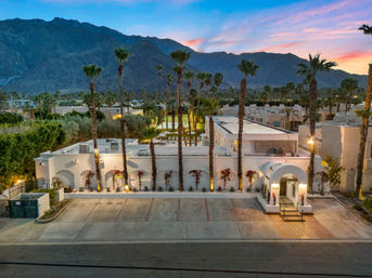 White stucco, Spanish-style villa with arched entry and palm-lined courtyard, warm evening lights and rugged desert mountains at sunset — palm tree resort vibe.