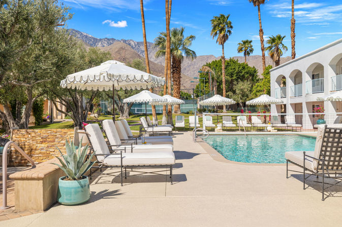 Sunny desert resort pool with white lounge chairs and scalloped umbrellas, palm trees and rugged mountains in the background.